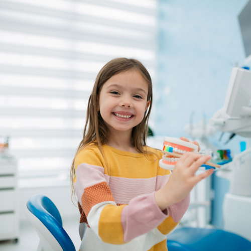Young girl holding colorful toothbrush in modern dental office setting, smiling at camera.