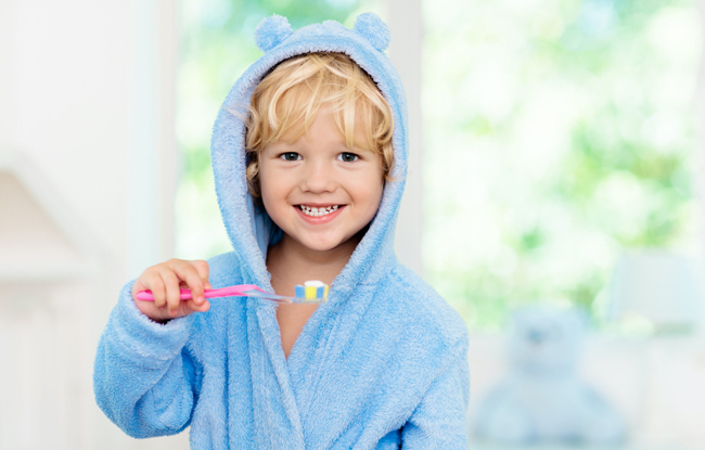 Happy young child in blue bathrobe holding toothbrush, smiling at camera in bright bathroom setting.