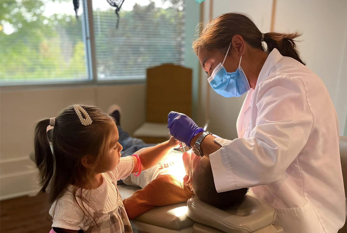 Pediatric dentist examining young girl's teeth in bright clinical treatment room with window view.