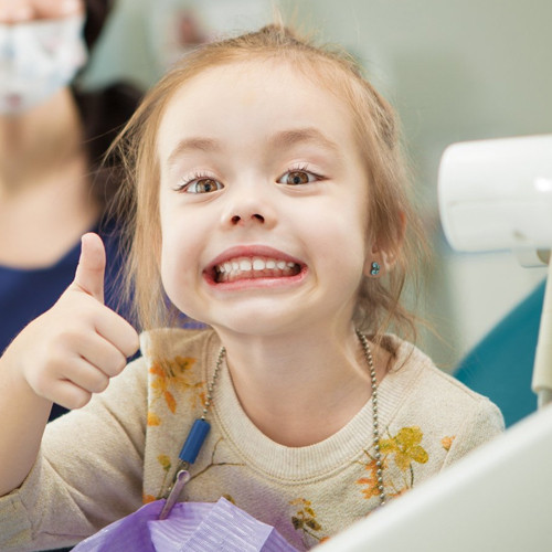 Young girl giving thumbs up smile in dental office setting, showing approval of pediatric dental care.
