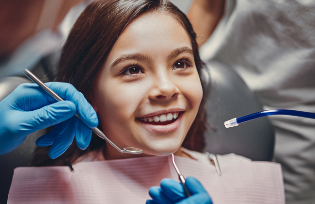 Young girl smiling at dental office while dentist holds dental tools wearing blue gloves.
