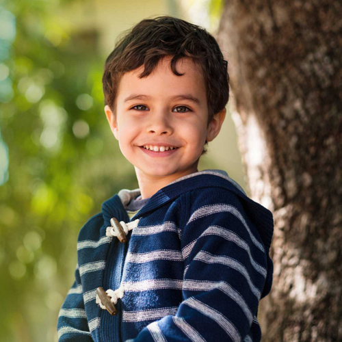 Young boy in striped blue jacket smiling outdoors by tree trunk.
