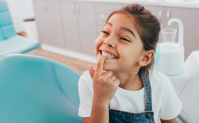 Young girl smiling while touching her teeth in a bright dental office with blue accents.