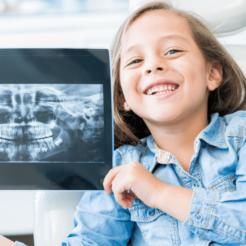 Young girl in denim shirt smiling while holding dental X-ray in bright clinical setting.