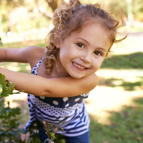 Happy young girl smiling outdoors in a blue striped shirt with arms raised.