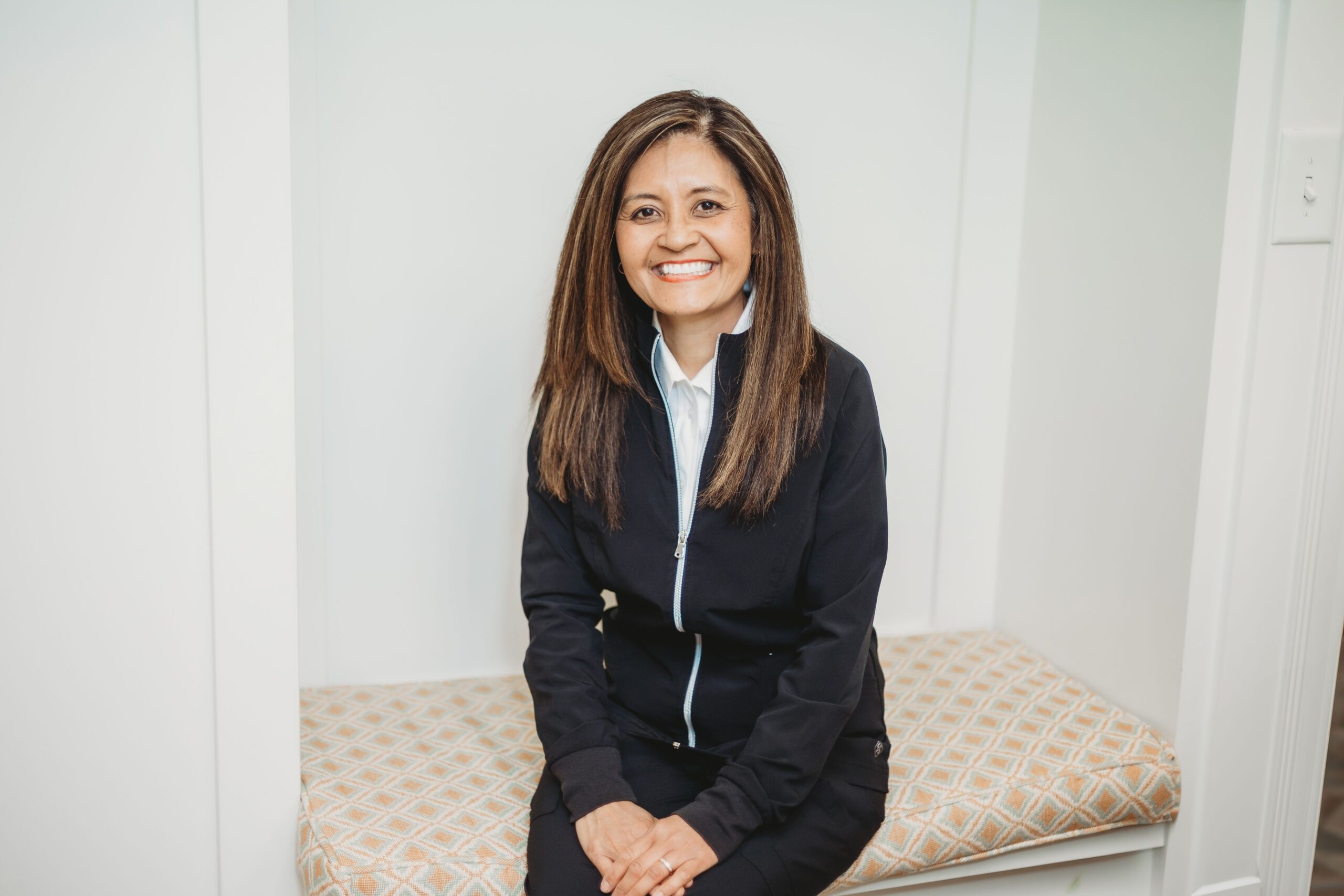 Professional woman in dark blazer sitting on dental chair, smiling at camera in clinical office setting.