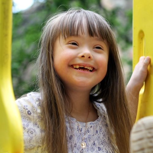 Young child with Down syndrome smiling outdoors near yellow playground equipment.
