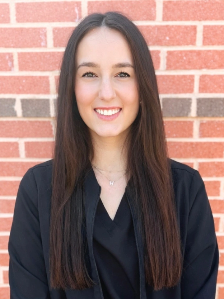 Professional headshot of woman in dark blazer against red brick wall background.