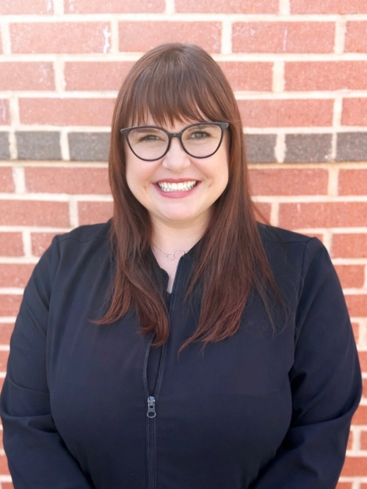 Woman with glasses smiling at camera against brick wall background, professional headshot.