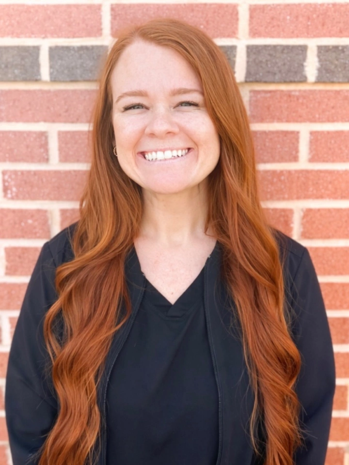 Woman with long red hair smiling at camera against red brick wall background.