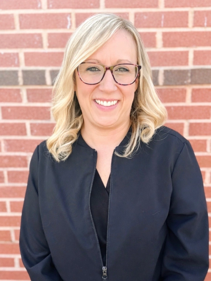 Professional headshot of woman with blonde hair and glasses against brick wall background.