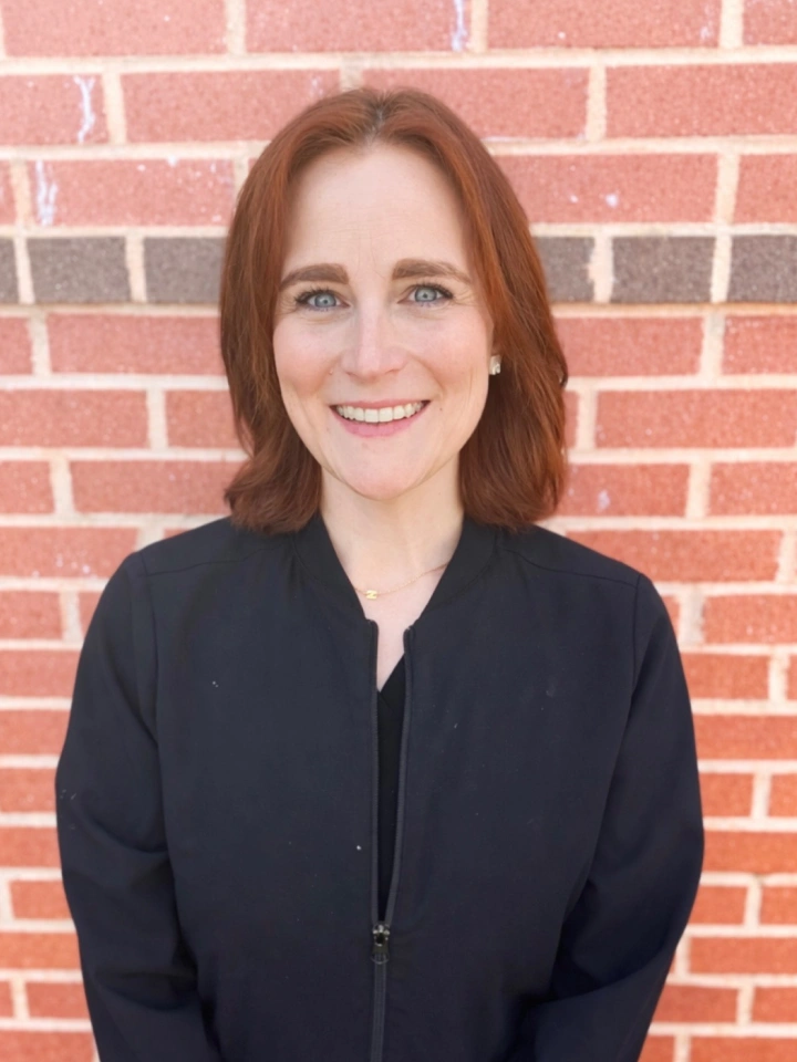 Professional headshot of woman with reddish-brown hair smiling against brick wall background.